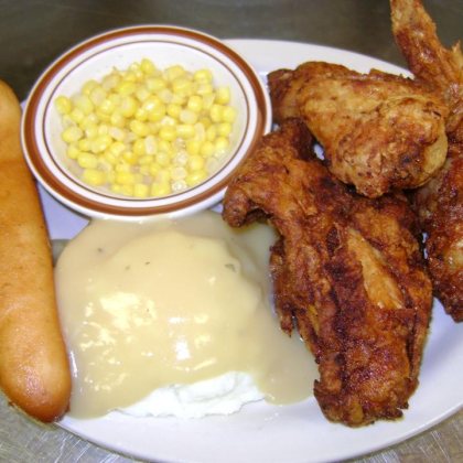 A plate of broasted chicken with mashed potatoes and gravy, sweetcorn in a side bowl and a breadstick.