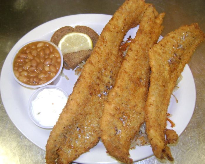 Three hand-breaded fish fillets over fried potatoes, with baked beans, homemade tartar sauce, dark rye bread and a lemon slice.