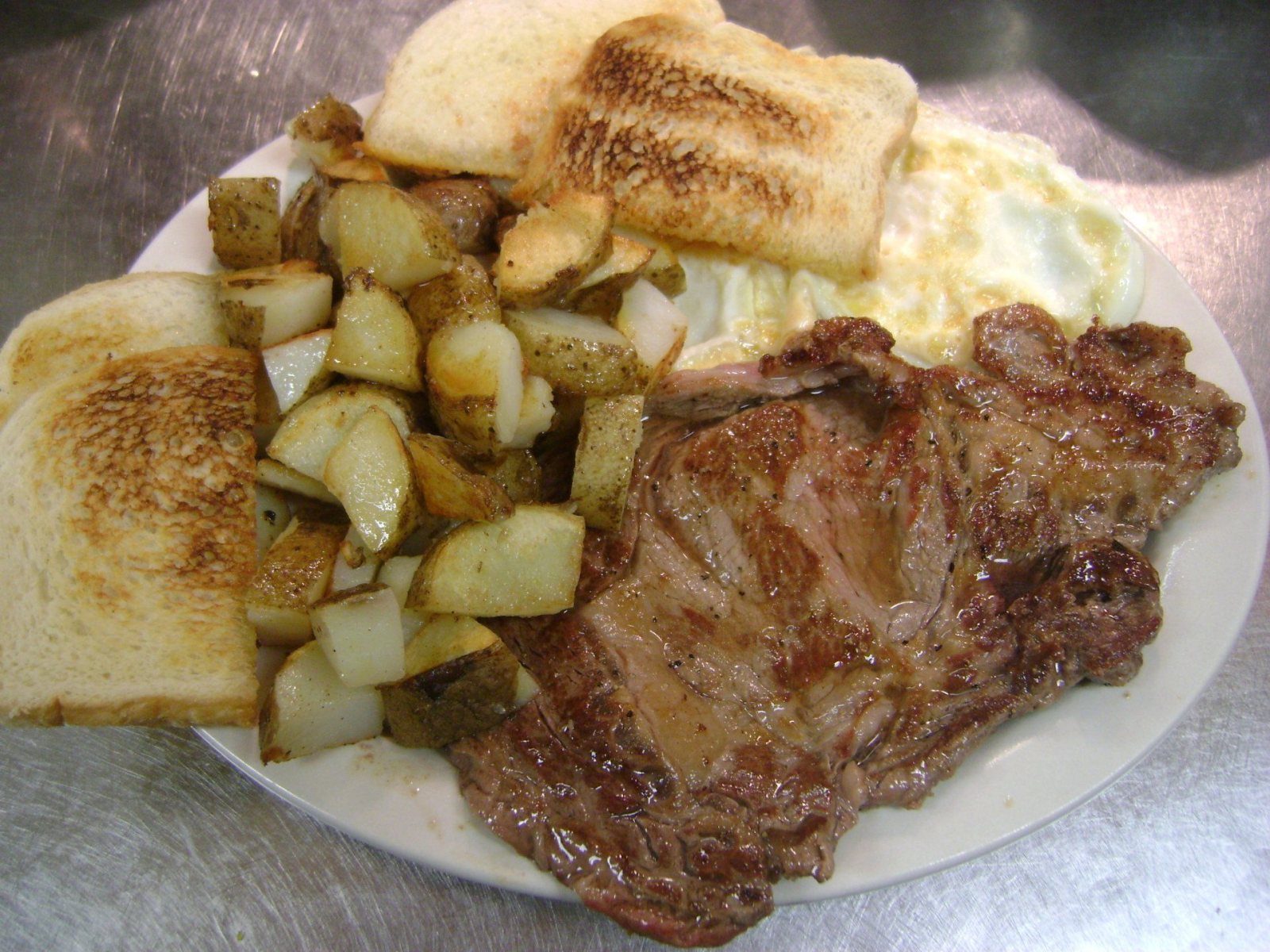 Steak and eggs: a ribeye, two fried eggs, fried taters and buttered toast on a white oval plate.