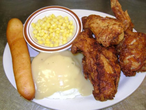 Broasted chicken with mashed potatoes and gravy, sweetcorn in a side bowl and a breadstick.