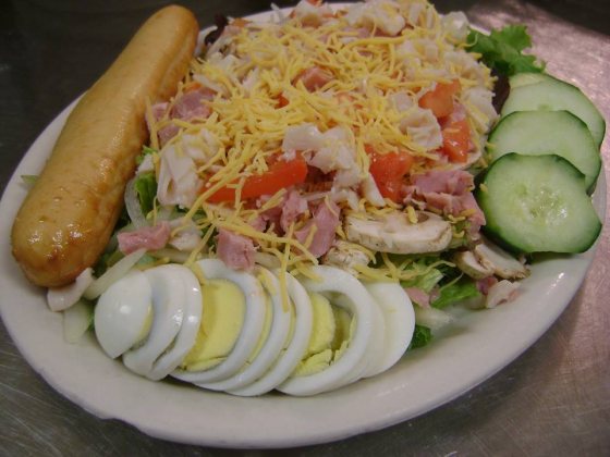A chef salad: shredded cheese, diced ham and turkey, tomato, mushrooms, cucumber, sliced hard-boiled egg and a breadstick.