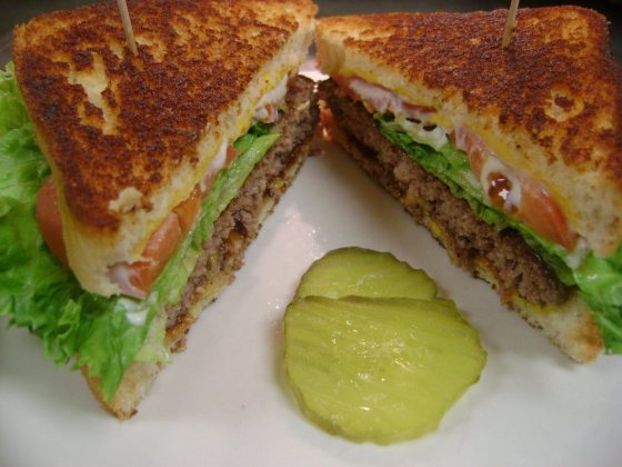 A burger on grilled Texas toast, cut in half, with lettuce, tomato, melted cheese and a pickle slice alongside.